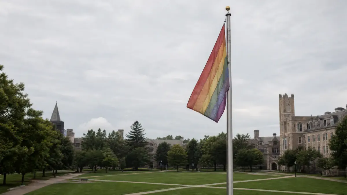 A faded rainbow Pride flag hanging on a college campus flagpole, with an empty quad in the background